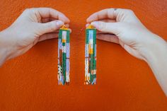 two hands are holding beaded earrings against an orange wall, one has green and white beads on it