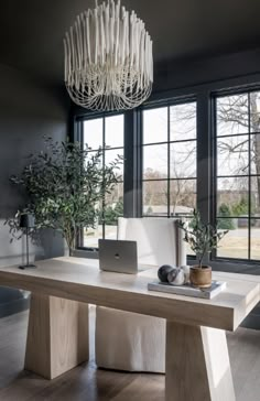 a table with a laptop on it in front of large windows and a chandelier