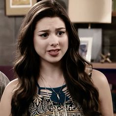 a woman with long brown hair wearing a top and necklace on her neck, standing in front of a table