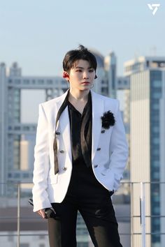 a young man in a white jacket and black shirt standing on a roof with his hands in his pockets