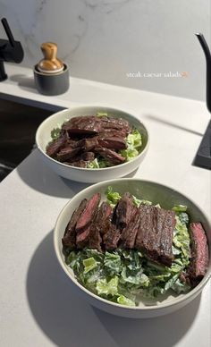 two white bowls filled with steak and lettuce on top of a kitchen counter