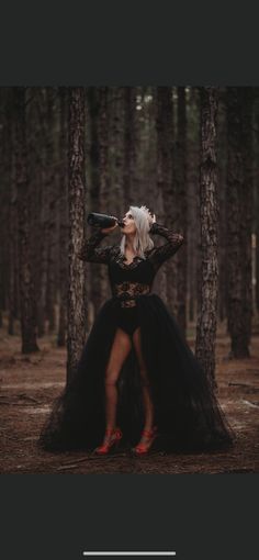 a woman with white hair and black dress standing in the woods holding a wine bottle