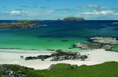 two people are standing on the beach looking out at the blue water and green land