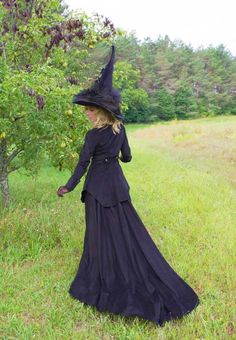 a woman in a long black dress and hat walking through the grass with her hand on her hip