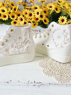 two pairs of white high top sneakers sitting next to yellow sunflowers on a table