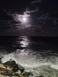 the full moon shines brightly over the ocean as waves crash on the rocks in front of it