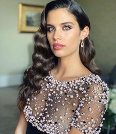 a woman with long dark hair wearing a black dress and pearls on her shoulders, looking at the camera