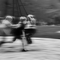 black and white photograph of two people riding on a swing set with blurry trees in the background