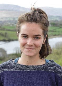 a woman standing in front of a river with hills and fields behind her, smiling at the camera