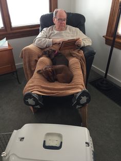 an old man sitting in a chair with his dog and looking at the book he is reading