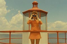 a woman in an orange dress is looking through binoculars at the ocean while standing on a boat