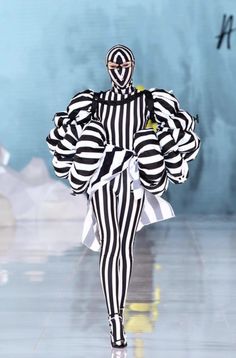 a model walks down the runway wearing black and white striped clothing with an unusual headpiece