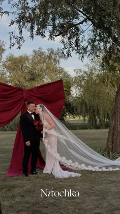 a bride and groom standing under a tree in front of a red drapeed curtain