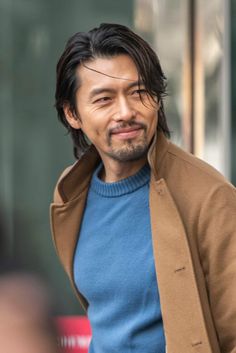 a man with long hair wearing a blue sweater and brown jacket is walking down the street