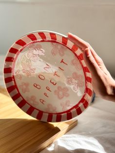 a person holding a red and white plate on top of a wooden table next to a window