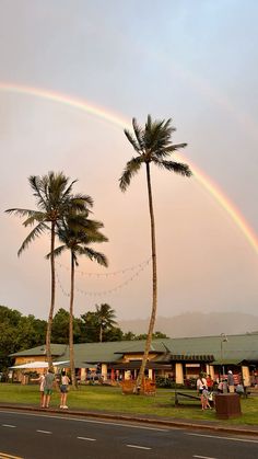 two palm trees with a rainbow in the sky above them and people standing on the grass