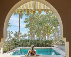 a woman sitting on the edge of a pool in front of an open archway with palm trees