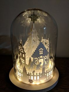 a glass dome with paper cut houses and trees in the inside, on a wooden table