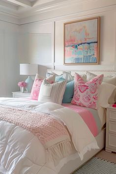 a white bed with pink and blue pillows in a bedroom next to a painting on the wall