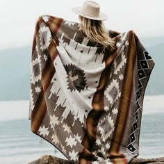 a woman wrapped in a blanket sitting on top of a rock next to the ocean