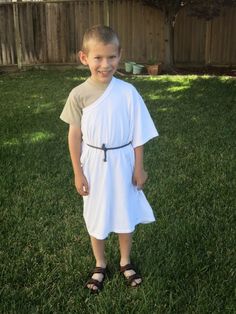 a young boy standing in the grass wearing a white dress with black straps on it