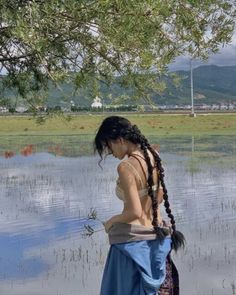 a woman with braids standing in the water next to a tree and looking down