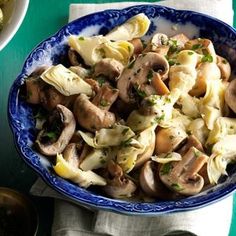 a blue bowl filled with pasta and mushrooms on top of a green tablecloth next to silverware