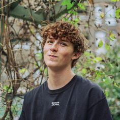 a young man with curly hair standing in front of a tree and looking up at the sky