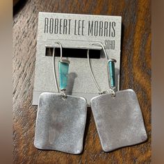 two square shaped silver earrings with turquoise stone accents on top of a wooden table next to a card