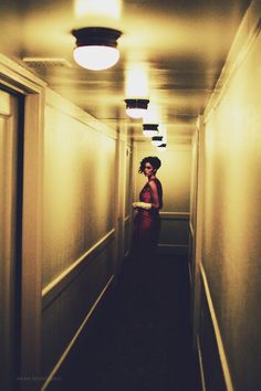 a woman in a red dress is walking down an empty hallway with lights on the ceiling