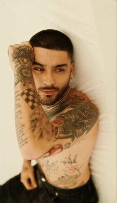 a man with tattoos on his arms and chest laying in bed next to a white wall