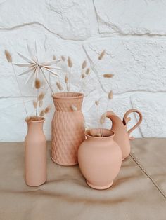 three pink vases with dried flowers in them on a brown tablecloth next to a white brick wall