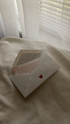 an open envelope with a heart on it sitting on a bed in front of blinds