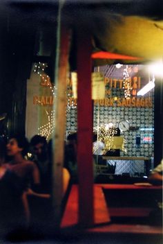 people are sitting at tables in front of a store window with lights on the windows