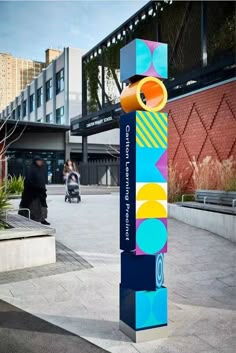 a tall colorful sign sitting on the side of a road next to a building with people walking by