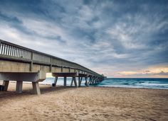 a long wooden pier sitting on top of a sandy beach next to the ocean under a cloudy sky