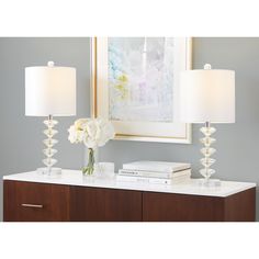 two lamps sitting on top of a white table next to a framed art piece and flowers