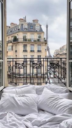 a bed with white sheets and pillows is in front of an open window overlooking the eiffel tower