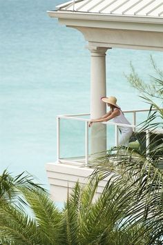 a woman wearing a hat leaning on a railing near the ocean with palm trees in front of her