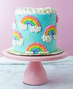 a blue cake decorated with rainbows and clouds on a pink stand in front of a pink wall