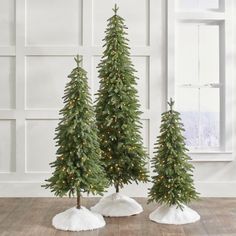 three artificial christmas trees with lights on them in front of a white wall and wooden floor