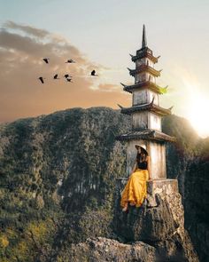 a woman sitting on top of a rock next to a tall tower with birds flying over it