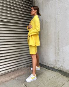 Fashion 2024 shines bright with this vibrant, sunshine-yellow ensemble, embodying the joyous spirit of spring and summer streetwear. The woman stands poised, her chic blazer and shorts set offering a playful yet polished look, cinched at the waist for a flattering silhouette. Paired with classic white sneakers that feature a pop of yellow, she presents a harmonious blend of casual comfort and style. Her sleek sunglasses and pulled-back hair accentuate the clean lines and fresh vibe of the outfit. This is a statement of bold color and relaxed tailoring, showcasing how Outfits 2024 celebrates personal style with energetic hues and effortless grace. Blazer Shorts Outfit, Athleisure Trend, Chic Blazer, Summer Streetwear, The Embrace, Classic Suit, Blazer And Shorts, Street Style Chic