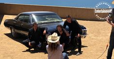 a group of people standing next to a black car on top of a dirt field