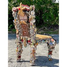 a bench made out of old objects in the middle of a parking lot with trees and bushes behind it