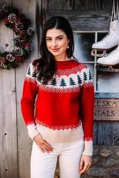 a woman standing in front of a wooden wall wearing a red sweater and white pants