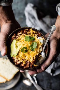 a person holding a bowl of chili with cheese and avocado on top next to some crackers