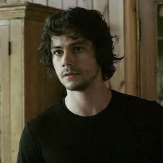 a man with curly hair and black shirt looking at the camera while standing in front of a cabinet