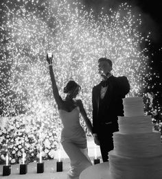 a bride and groom standing next to a wedding cake with fireworks in the background at night