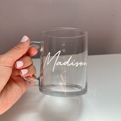 a hand holding a glass mug with the word madison on it and white writing in cursive font